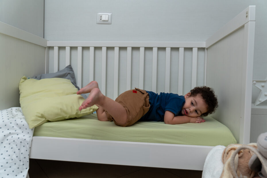 Un bambino sta sdraiato su un letto con sponde bianche, con un cuscino verde chiaro e una coperta a pois sul bordo.