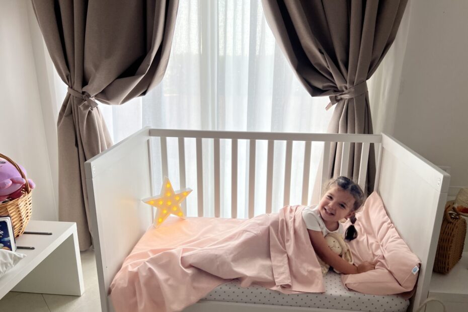 Una bambina sorridente è sdraiata in un lettino bianco con lenzuola rosa e abbraccia un peluche. La stanza è luminosa grazie alla luce naturale che filtra attraverso tende marroni. Accanto al lettino c'è una lampada a forma di stella illuminata e un cesto di vimini con giocattoli.