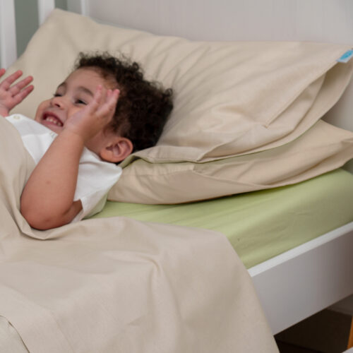 Un bambino sorride felicemente mentre è sdraiato su un lettino, con due cuscini avorio chiaro dietro la testa. È coperto da un lenzuolo coordinato dello stesso colore. Il materasso è rivestito da un lenzuolo verde chiaro, creando un contrasto morbido e rilassante. Il bambino ha un atteggiamento giocoso, trasmettendo un'atmosfera accogliente e serena.