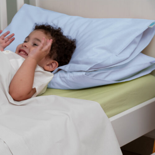 Bambino sorridente sdraiato su un lettino, con due cuscini di colore azzurro chiaro sotto la testa. È coperto da un lenzuolo bianco, mentre il materasso è rivestito con un lenzuolo verde. Il bambino sembra giocoso e rilassato, creando un'atmosfera allegra e serena.