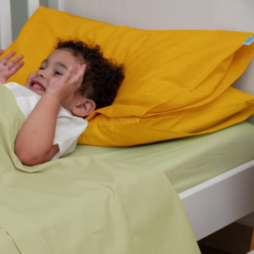 Bambino sorridente sdraiato su un lettino con lenzuola verde chiaro e un cuscino doppio sotto la testa di colore giallo. Il materasso è coperto da un lenzuolo verde chiaro. Il bambino sembra felice e rilassato, immerso in un momento di gioco o riposo.