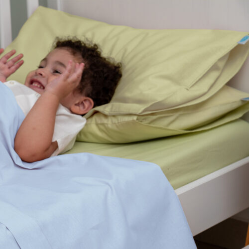 Bambino sorridente sdraiato su un lettino, con due cuscini di colore verde chiaro sotto la testa. È coperto da un lenzuolo azzurro chiaro, mentre il materasso è rivestito con un lenzuolo verde. Il bambino sembra giocoso e rilassato, creando un'atmosfera allegra e serena.