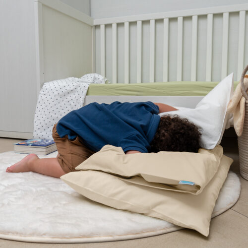 Un bambino gioca disteso su un tappeto soffice bianco, appoggiato a due cuscini avorio chiaro. Sembra stia esplorando o riposandosi, creando un'atmosfera rilassata. Sullo sfondo si vede un lettino con un materasso verde chiaro e un libro appoggiato a terra. L'ambiente appare tranquillo e accogliente, con una combinazione di colori chiari e pastello.