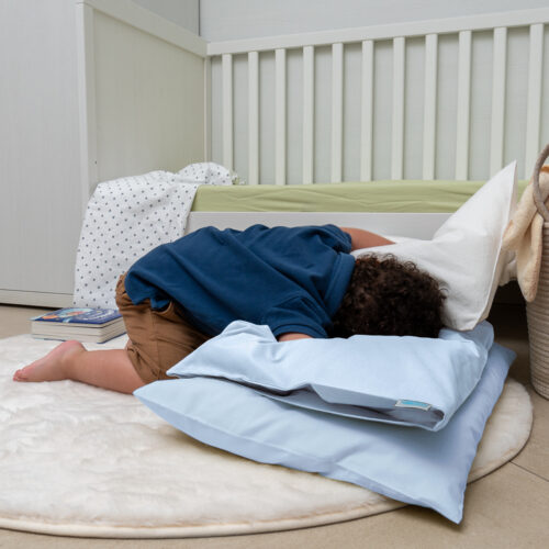 Bambino sdraiato a pancia in giù su un tappeto bianco, appoggiato su due cuscini, uno bianco e uno azzurro chiaro. Indossa una maglietta blu e pantaloncini marroni. Accanto al lettino si vedono un libro e un lenzuolo a pois. L'atmosfera è rilassata e giocosa.