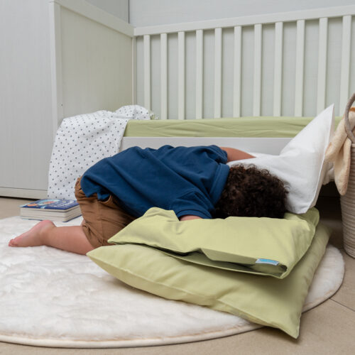 Bambino sdraiato a pancia in giù su un tappeto bianco soffice, appoggiato su cuscini verdi chiaro. Indossa una maglietta blu e pantaloncini marroni. Sullo sfondo c'è un lettino con lenzuolo verde chiaro, e un libro è posato a terra accanto a un lenzuolo a pois.