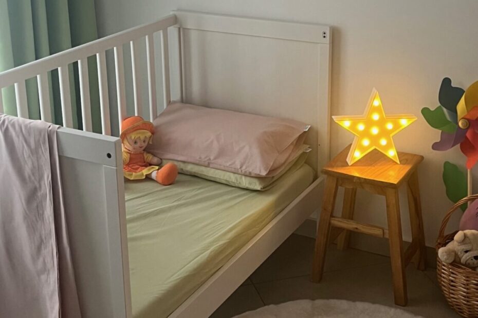 Una cameretta per bambini con una culla bianca, decorata con una ghirlanda colorata e una stella luminosa accesa su uno sgabello di legno. Accanto al letto c'è una bambola di pezza arancione e un cesto con peluche.