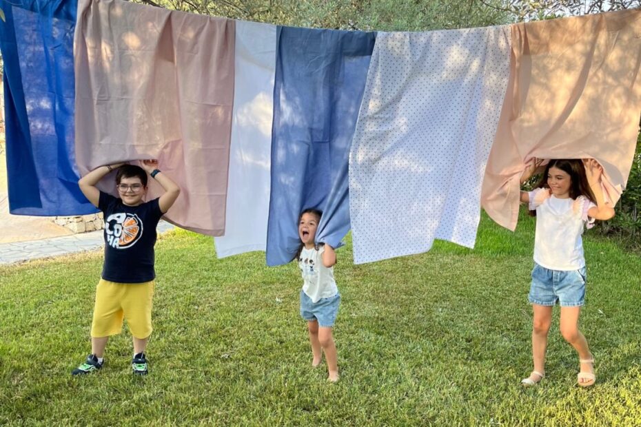 Tre bambini giocano all'aperto sotto delle lenzuola appese, sollevandole sopra le loro teste.