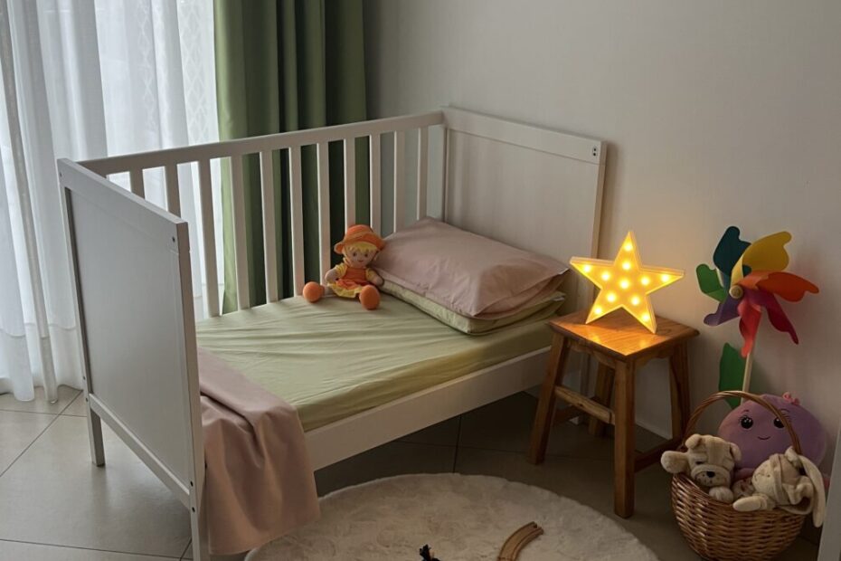 Camera da letto per bambini con lettino bianco e lenzuola con colori verde e rosa, giocattoli, una luce a forma di stella e una cesta piena di peluche.