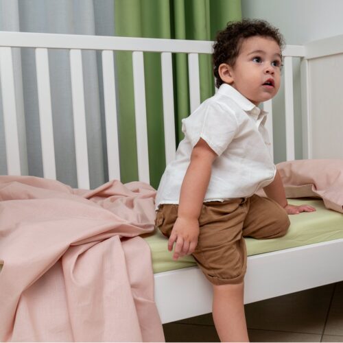 Bambino seduto in una culla bianca, vestito con una camicia bianca e pantaloncini marroni. Guarda verso l'alto con un'espressione curiosa. Sulla culla c'è un lenzuolo rosa sepia e sullo sfondo sono visibili delle tende verdi e bianche.