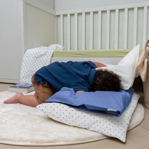 Un bambino gioca disteso su un tappeto soffice bianco, appoggiato a due cuscini uno bianco con pois blue e uno blue chiaro. Sembra stia esplorando o riposandosi, creando un'atmosfera rilassata. Sullo sfondo si vede un lettino con un materasso verde chiaro e un libro appoggiato a terra. L'ambiente appare tranquillo e accogliente, con una combinazione di colori chiari e pastello.