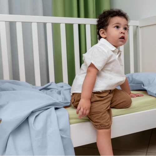 Bambino seduto in una culla bianca, vestito con una camicia bianca e pantaloncini marroni. Guarda verso l'alto con un'espressione curiosa. Sulla culla c'è un lenzuolo azzurro e sullo sfondo sono visibili delle tende verdi e bianche.