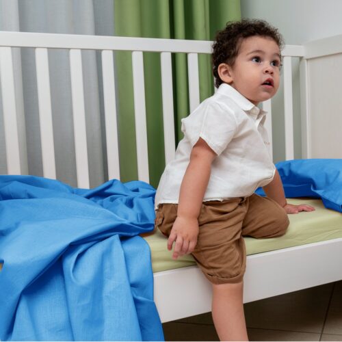 Bambino seduto in una culla bianca, vestito con una camicia bianca e pantaloncini marroni. Guarda verso l'alto con un'espressione curiosa. Sulla culla c'è un lenzuolo blu e sullo sfondo sono visibili delle tende verdi e bianche.