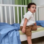 Bambino seduto in una culla bianca, vestito con una camicia bianca e pantaloncini marroni. Guarda verso l'alto con un'espressione curiosa. Sulla culla c'è un lenzuolo blue smoke e sullo sfondo sono visibili delle tende verdi e bianche.