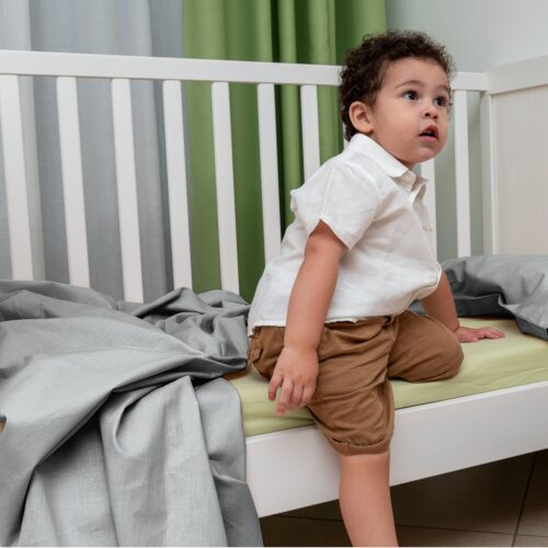 Bambino seduto in una culla bianca, vestito con una camicia bianca e pantaloncini marroni. Guarda verso l'alto con un'espressione curiosa. Sulla culla c'è un lenzuolo grigio e sullo sfondo sono visibili delle tende verdi e bianche.