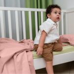 Bambino seduto in una culla bianca, vestito con una camicia bianca e pantaloncini marroni. Guarda verso l'alto con un'espressione curiosa. Sulla culla c'è un lenzuolo rosa praline e sullo sfondo sono visibili delle tende verdi e bianche.