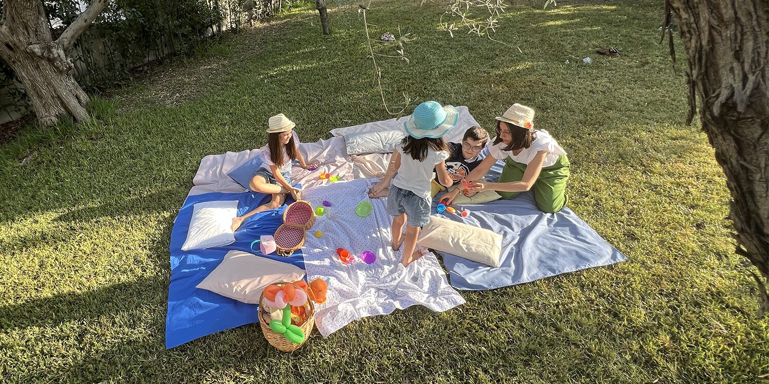 Un gruppo di bambini e adulti seduti su delle lenzuola di diverso colore stesi sull'erba, immersi in un momento di gioco e relax all'aperto. I bambini giocano con palline colorate e cesti, mentre indossano cappelli di paglia. La scena trasmette allegria e tranquillità in un ambiente verde e soleggiato.