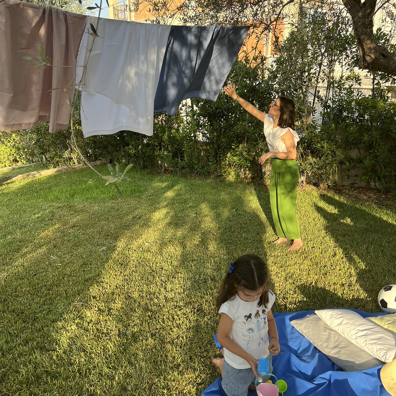 Bambina che gioca sull'erba accanto a un lenzuolo blu, mentre una donna osserva delle lenzuola colorate appese, mosse dal vento. La scena si svolge in un giardino soleggiato, con un'atmosfera tranquilla e rilassante in mezzo alla natura.